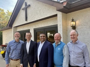 A group of smiling men standing outside Windsor Chapel, a welcoming Christian church with a cross on the building.